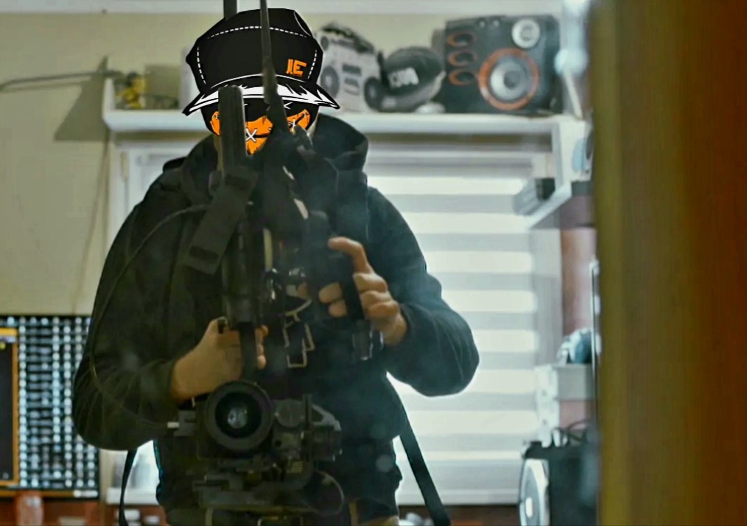 Operator kamery z charakterystycznym filtrem graficznym na twarzy i czapce, trzymający profesjonalny sprzęt filmowy w pomieszczeniu z widocznym sprzętem audio na półce.