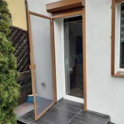 Otwarta moskitiera ramkowa w kolorze brązowym, zamontowana przy drzwiach balkonowych z roletą zewnętrzną, widoczna szczotka do zamiatania na ciemnych płytkach podłogowych.