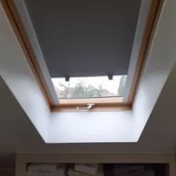 Szara roleta zamontowana w oknie dachowym z drewnianą ramą, widok z dołu na tle nieba i zabudowań.