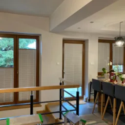 Jasne wnętrze domu z drewnianymi oknami, na których zamontowane są białe, poziome osłony okienne typu 'plaster miodu', częściowo zasłonięte, widoczne schody z drewnianą balustradą i fragment kuchni...