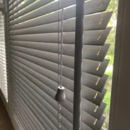 Szare żaluzje aluminiowe z widocznym mechanizmem sznurkowym i obciążnikiem, częściowo zasłonięte okno z zielenią na zewnątrz.