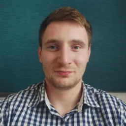 Portret młodego mężczyzny w koszuli w kratę na tle turkusowej ściany z ramkami, z delikatnym uśmiechem i lekkim zarostem.