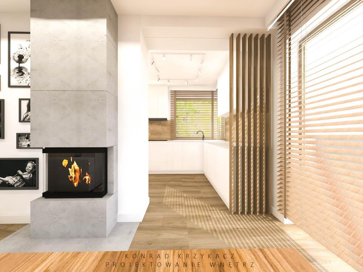 Wnętrze mieszkania z nowoczesnym kominkiem obłożonym szarym kamieniem, drewnianą podłogą i żaluzjami. W tle widoczna biała kuchnia z drewnianymi blatami.