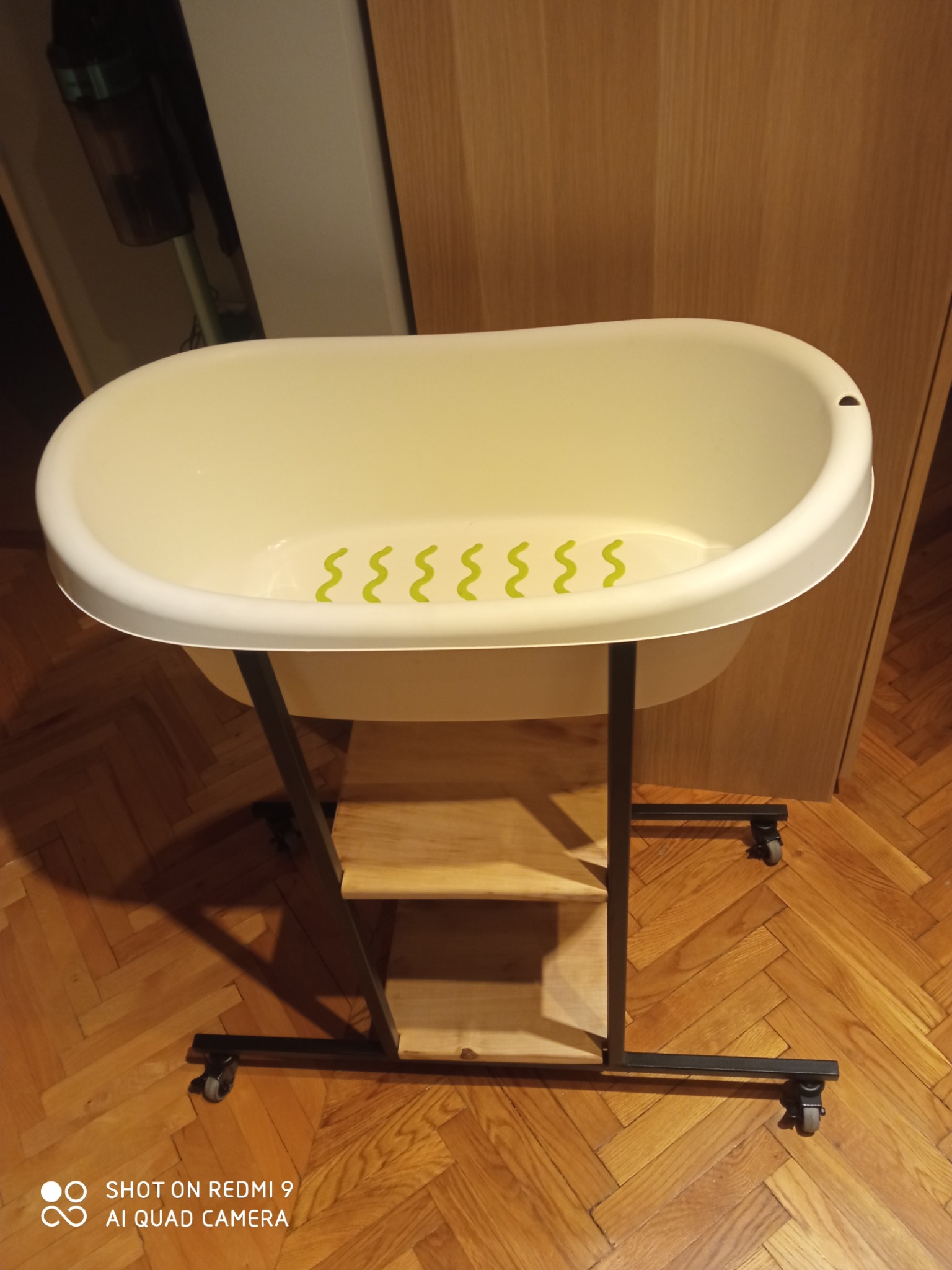 Biała wanna dla dziecka na metalowym stelażu z drewnianą półką, ustawiona na parkiecie w pomieszczeniu z drewnianymi meblami.