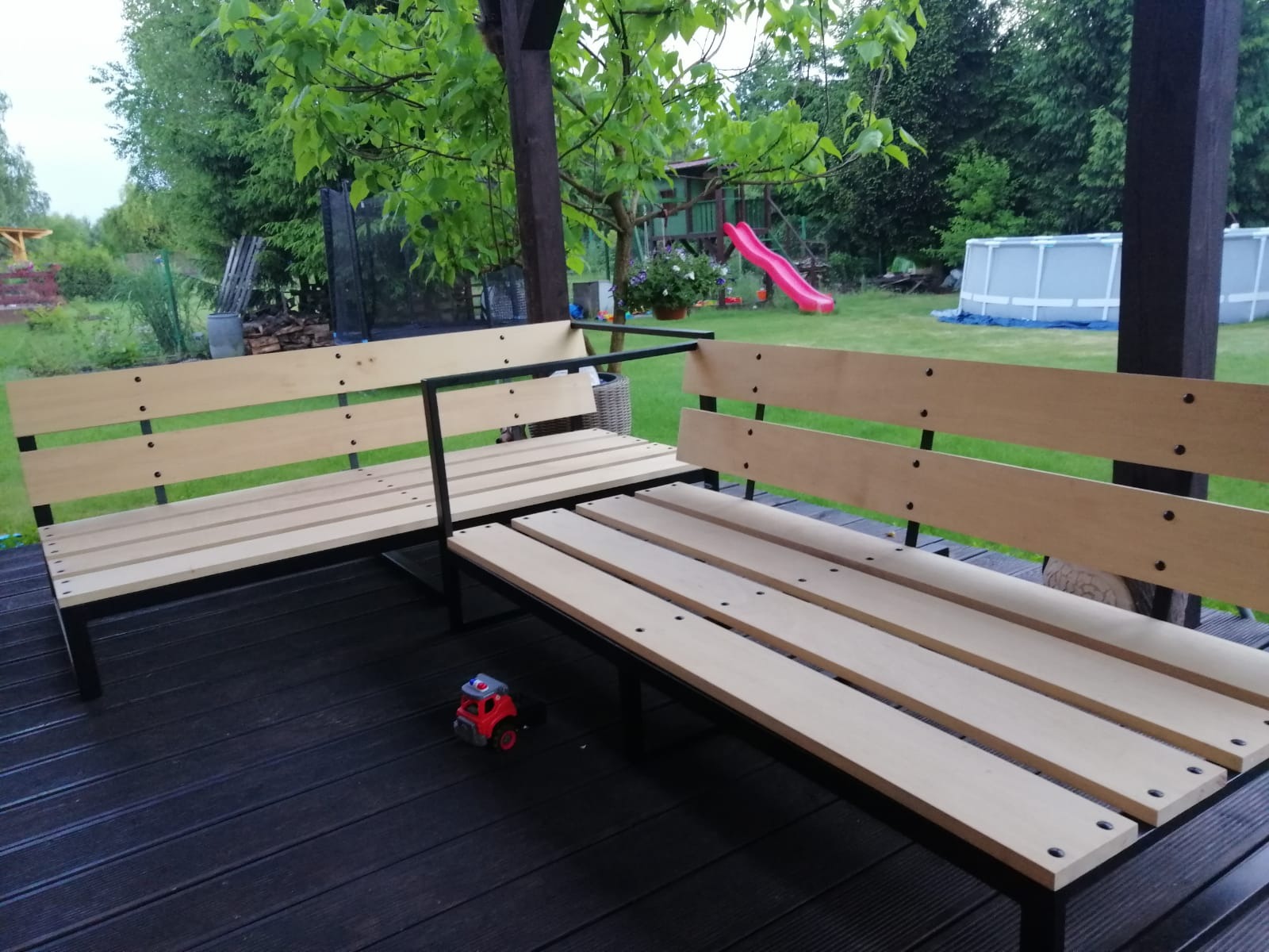 Kątowy zestaw mebli ogrodowych z jasnego drewna i czarnego metalu na ciemnym tarasie, z zabawką-autkiem w rogu; w tle trawnik z trampoliną i basenem.