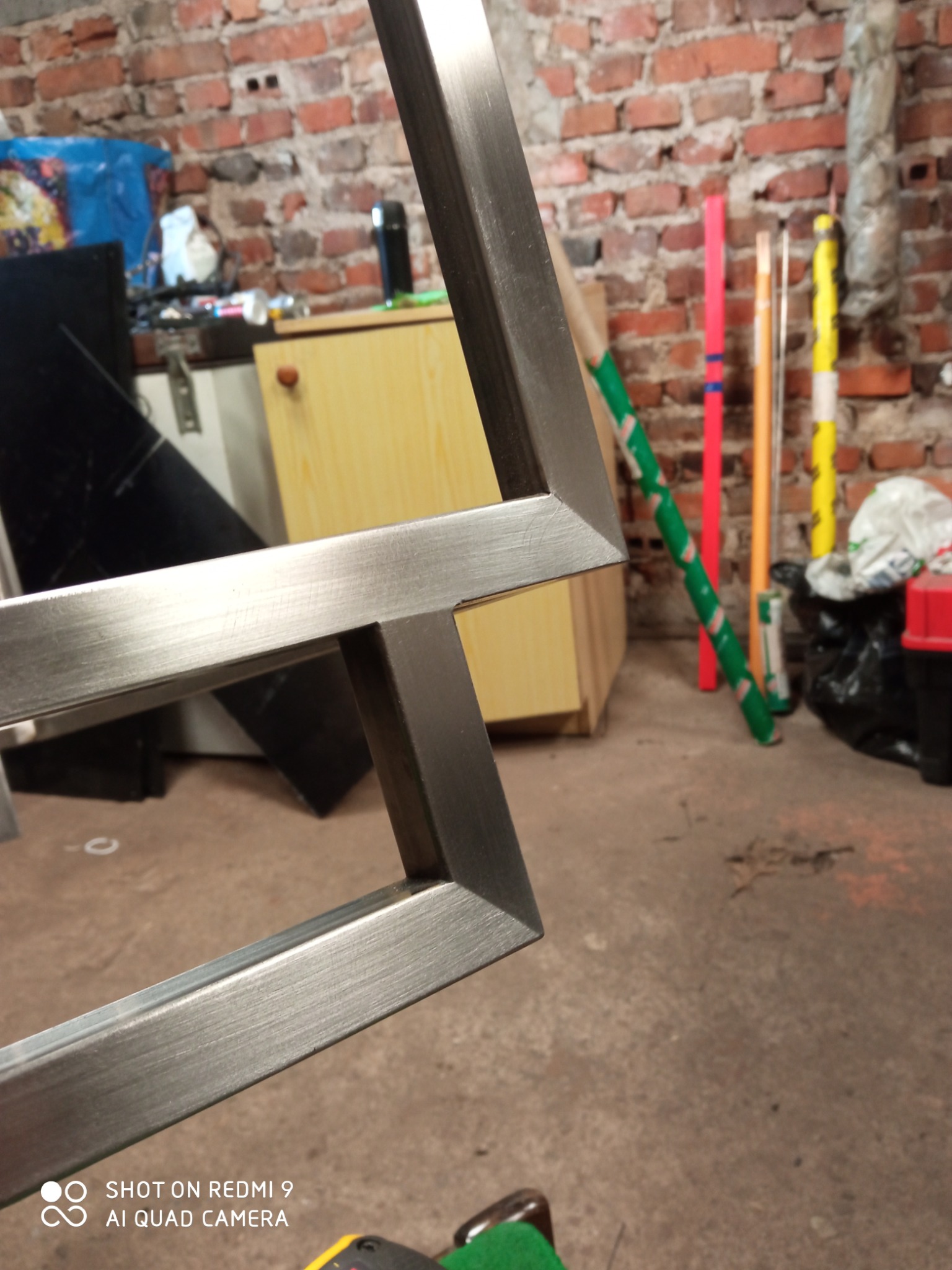 Fragment metalowej ramy spawanej z profili, widoczne spawy, w tle ceglana ściana i elementy wyposażenia warsztatu.