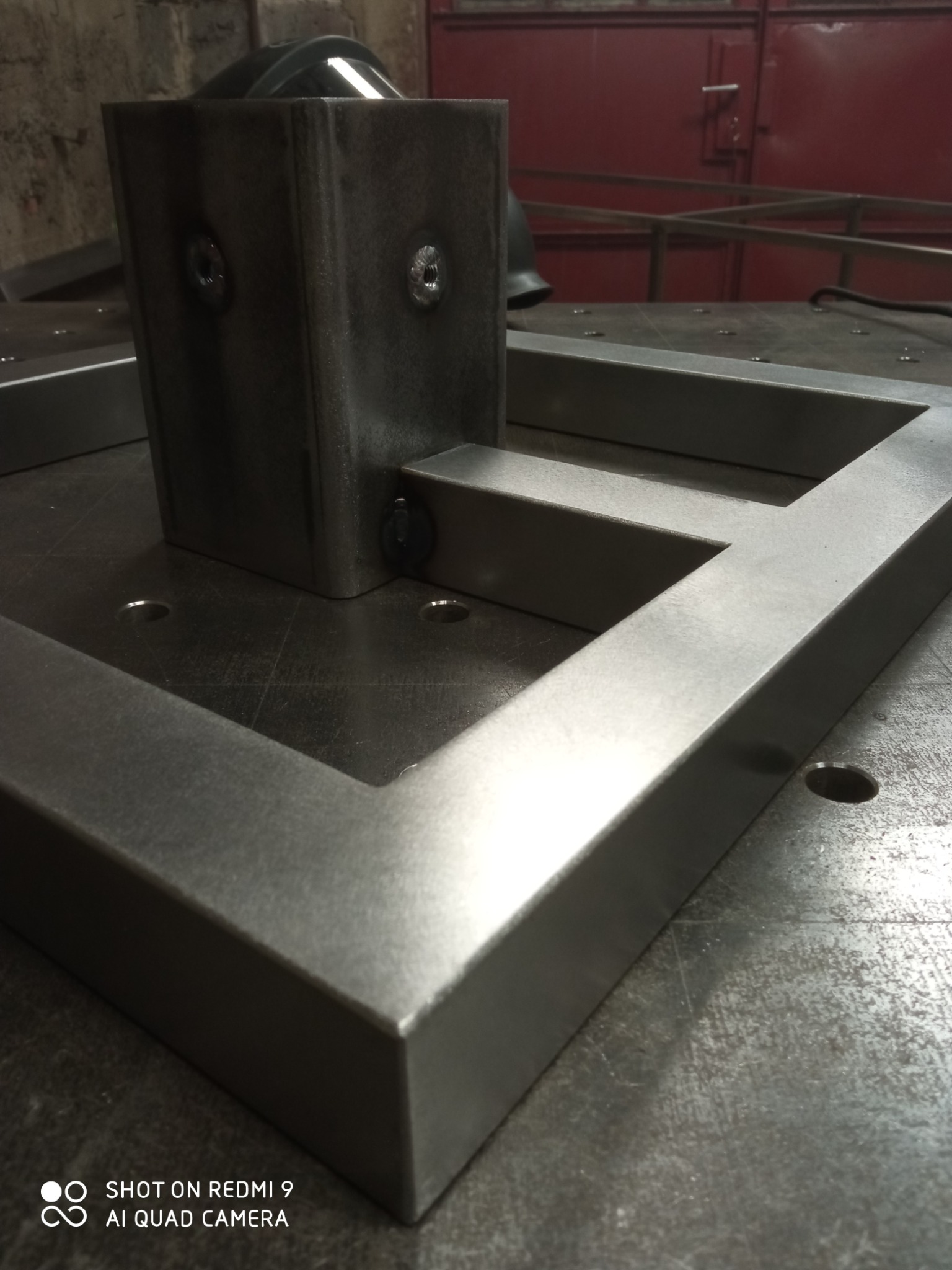 Metalowa konstrukcja spawana na stole spawalniczym, widoczne spawy punktowe łączące elementy, surowy metal z delikatnymi śladami obróbki.