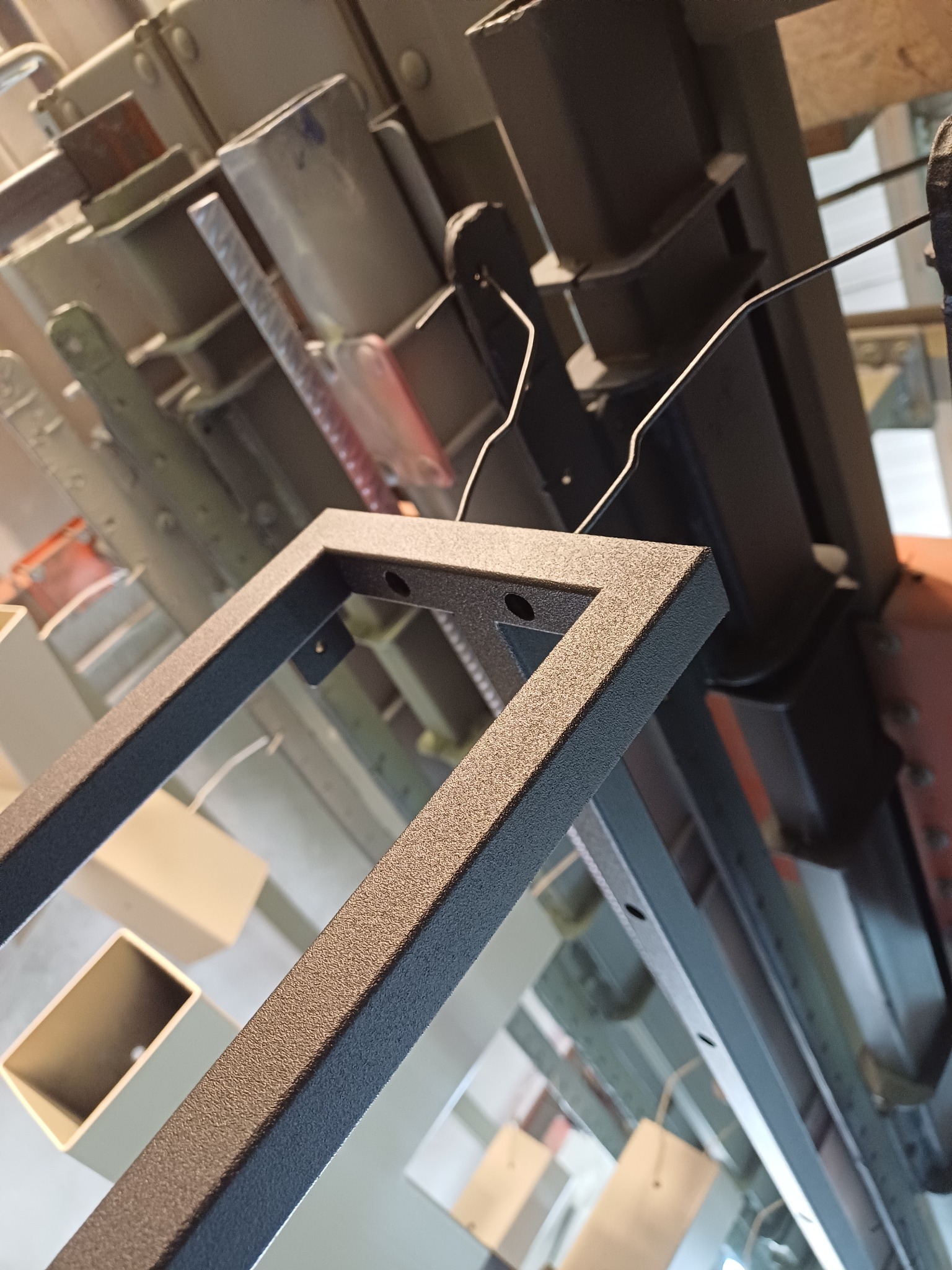 Metalowa rama pomalowana proszkowo, zawieszona na linii produkcyjnej w lakierni, widoczne otwory montażowe.