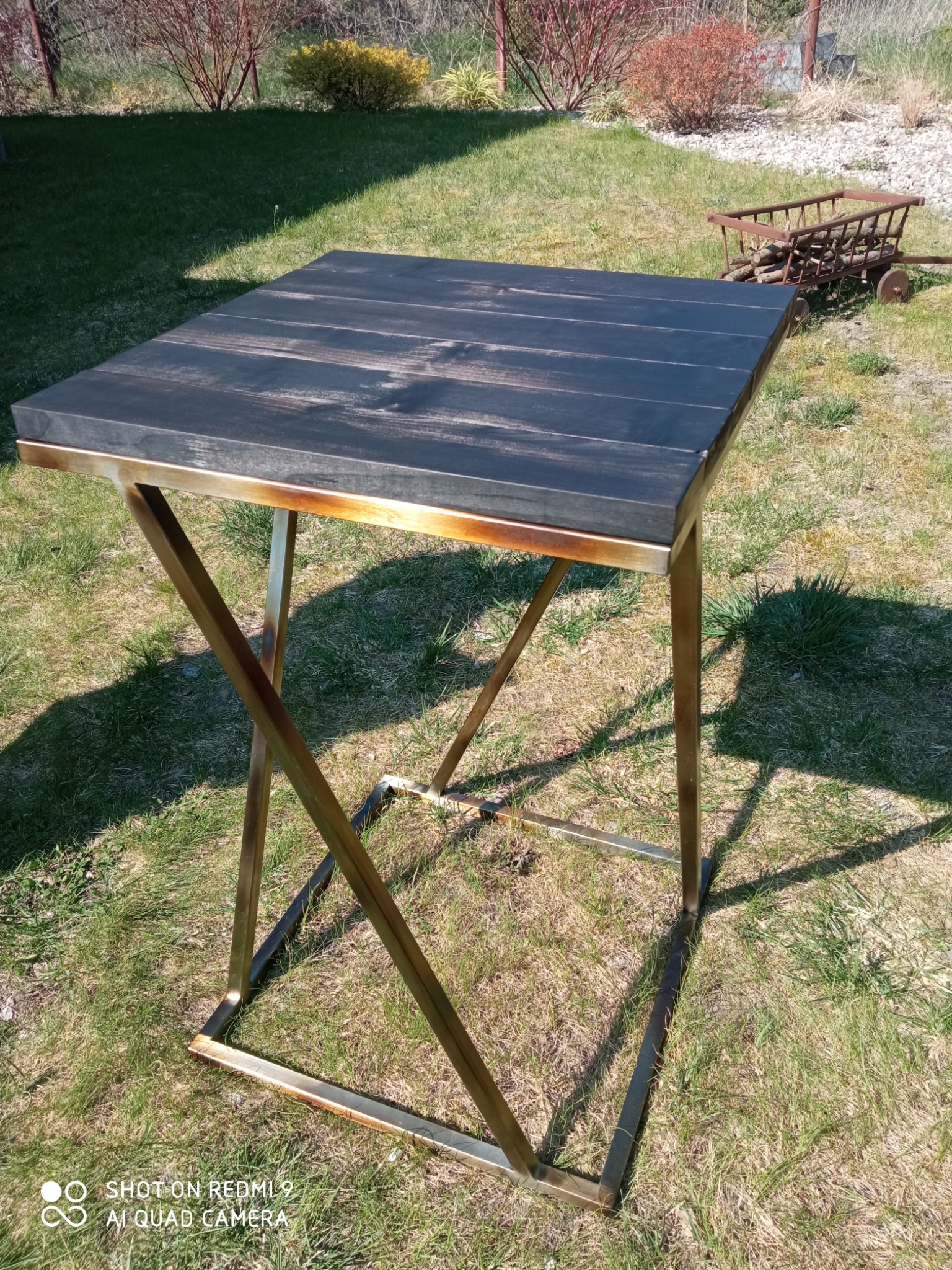 Mały, kwadratowy stolik z blatem z ciemnego drewna i metalową, spawaną ramą w kolorze złotym, stojący na trawie w ogrodzie; w tle dekoracyjny wózek z drewnem.