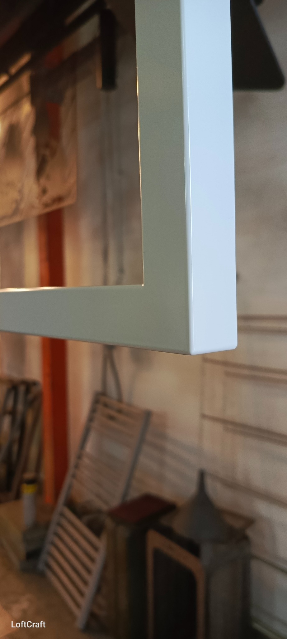 Biały, pomalowany proszkowo profil metalowy o przekroju kwadratowym, widoczny z bliska, w tle warsztat z rozłożonymi krzesłami i innymi elementami wyposażenia.