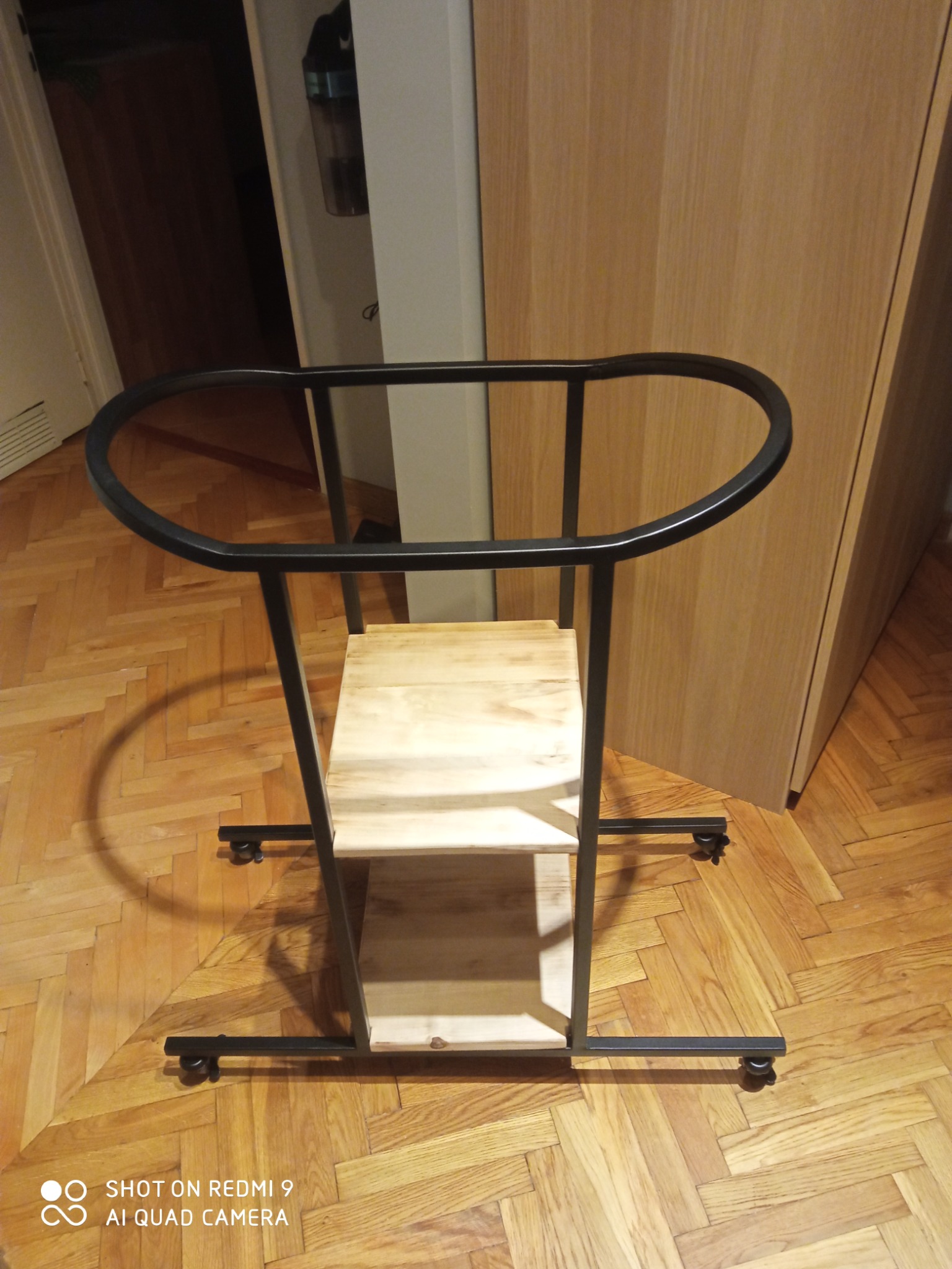 Metalowy wózek transportowy z drewnianymi półkami na kółkach, stojący na parkiecie ułożonym w jodełkę.