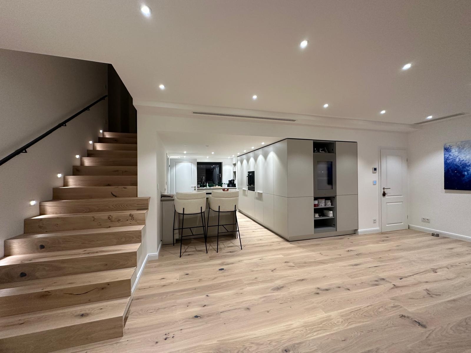 Nowoczesne wnętrze domu z drewnianymi schodami z podświetleniem, parkietem i zabudową kuchenną w odcieniach szarości. Minimalistyczny design, oświetlenie punktowe.