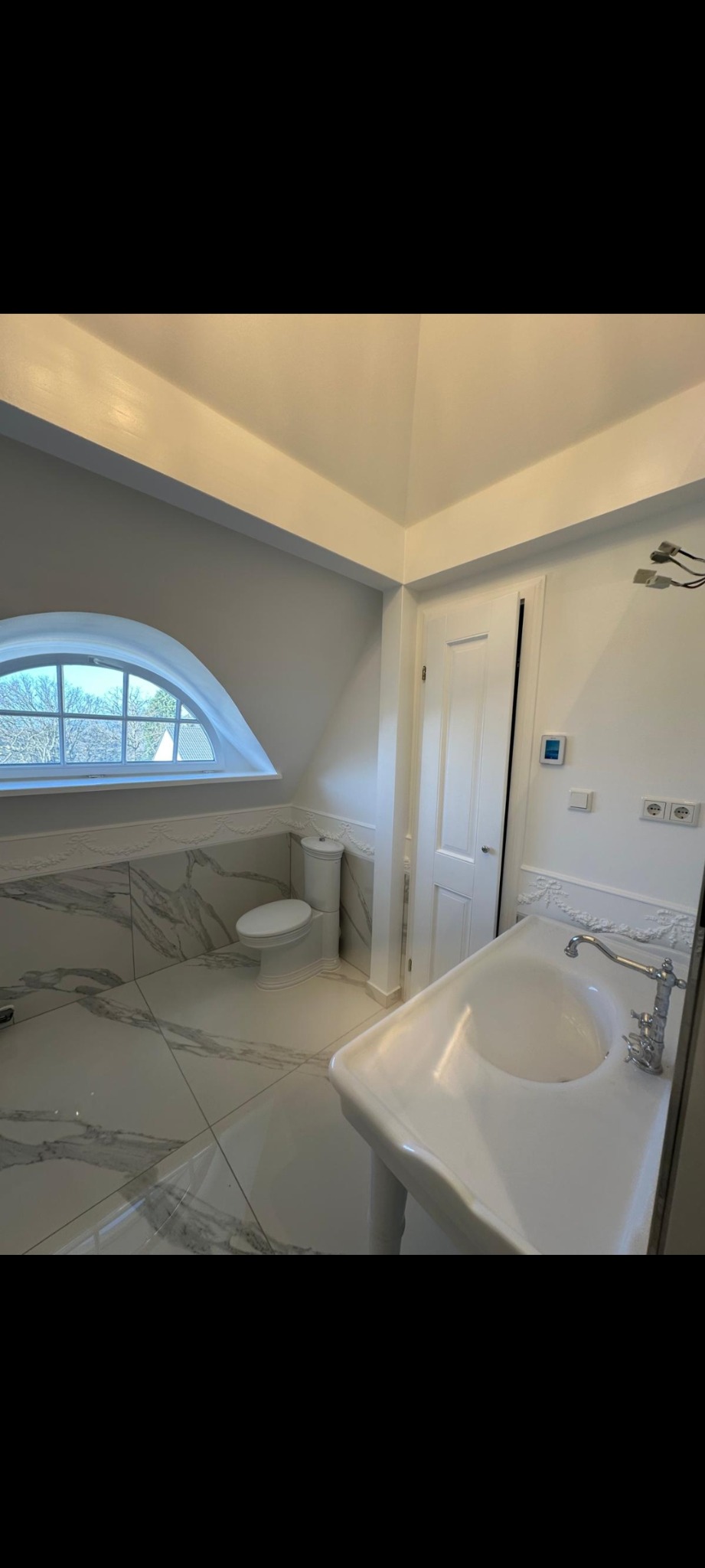 Jasna łazienka na poddaszu z marmurowymi płytkami, owalnym oknem i klasyczną umywalką na nodze. Białe ściany i drzwi. Widok na zewnątrz przez okno.