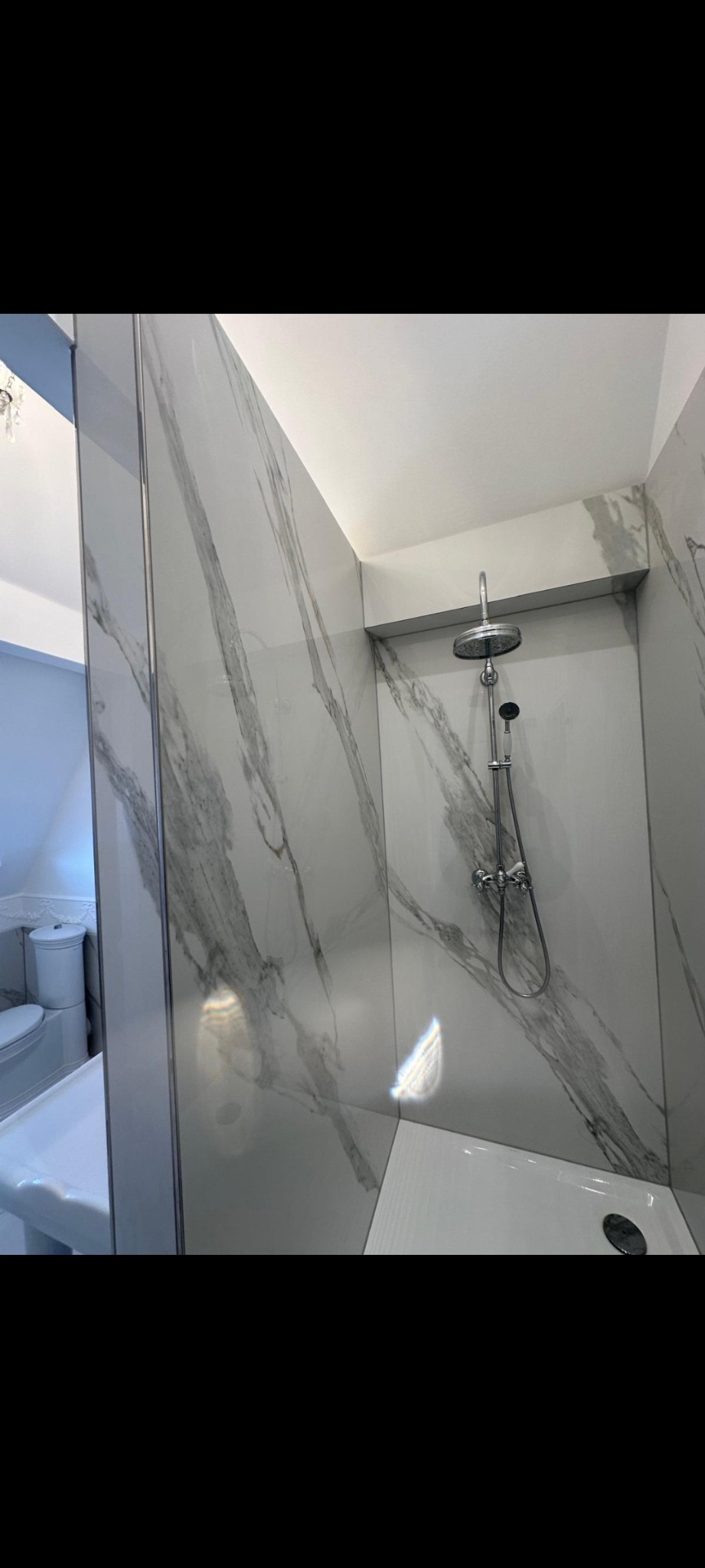 Nowoczesna kabina prysznicowa z marmurowymi płytkami i deszczownicą. Minimalistyczny design, jasne oświetlenie, chromowane wykończenia. Widok na umywalkę i toaletę.