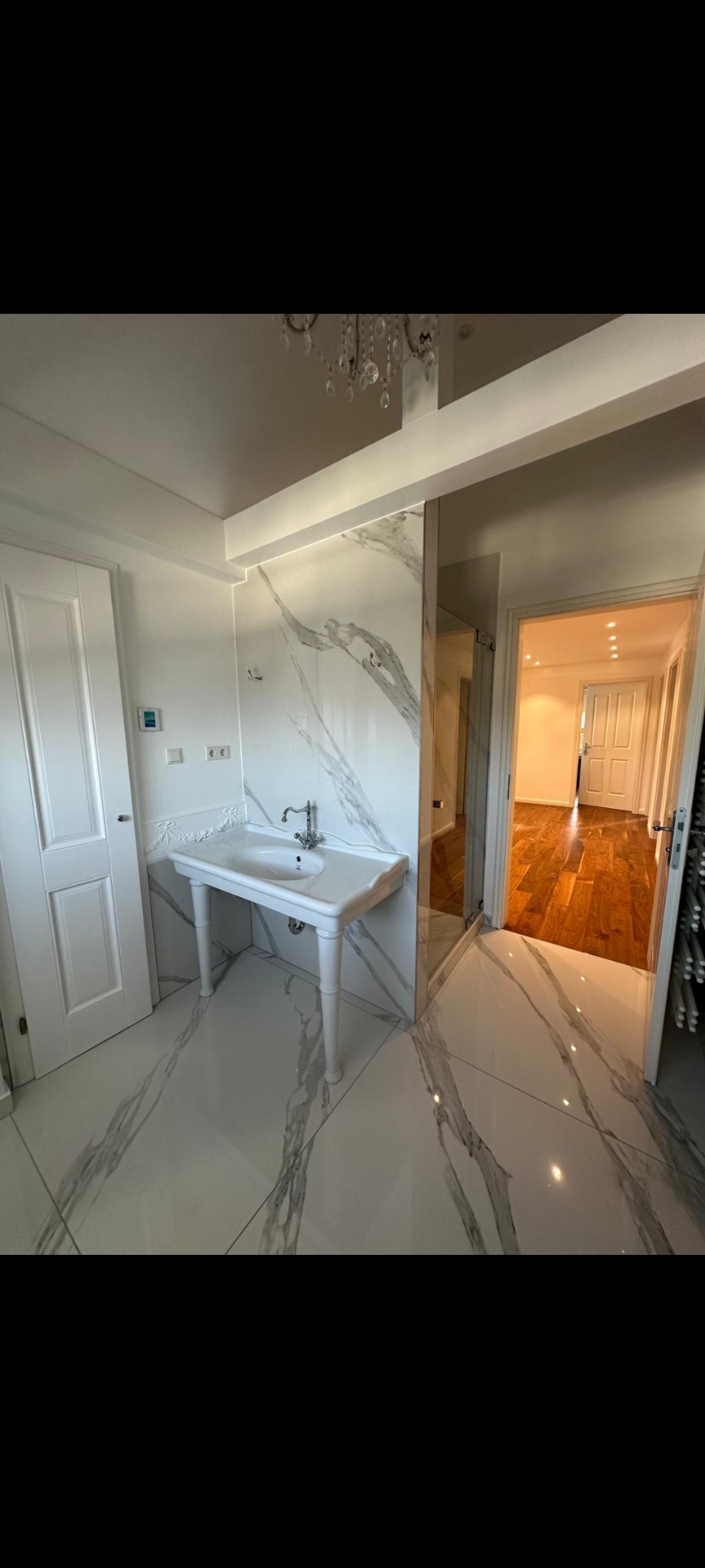 Elegancka łazienka z marmurowymi płytkami, wolnostojącą umywalką na nóżkach i kryształowym żyrandolem. Widok na sąsiednie pomieszczenie z drewnianą podłogą.