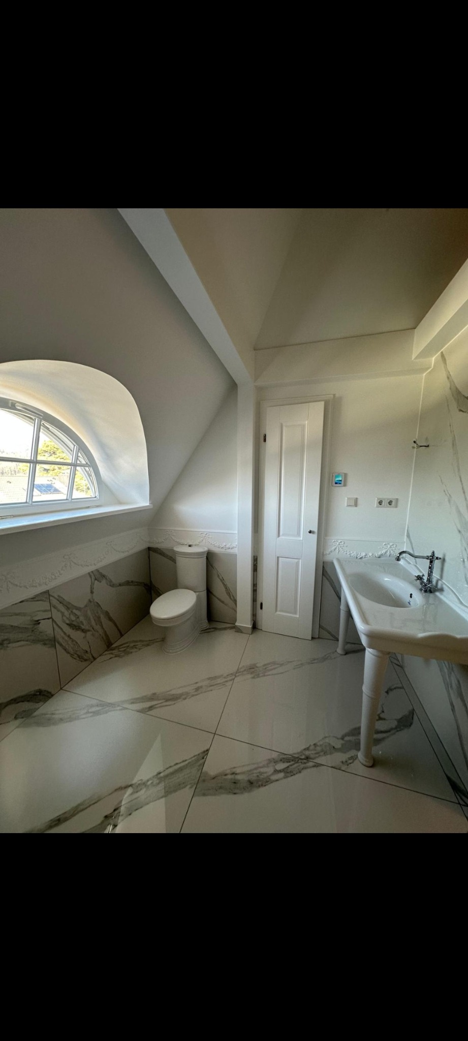 Jasna łazienka na poddaszu z oknem w kształcie półkola. Białe płytki imitujące marmur na podłodze i ścianach. Umywalka na ozdobnych nogach i toaleta w stylu retro.