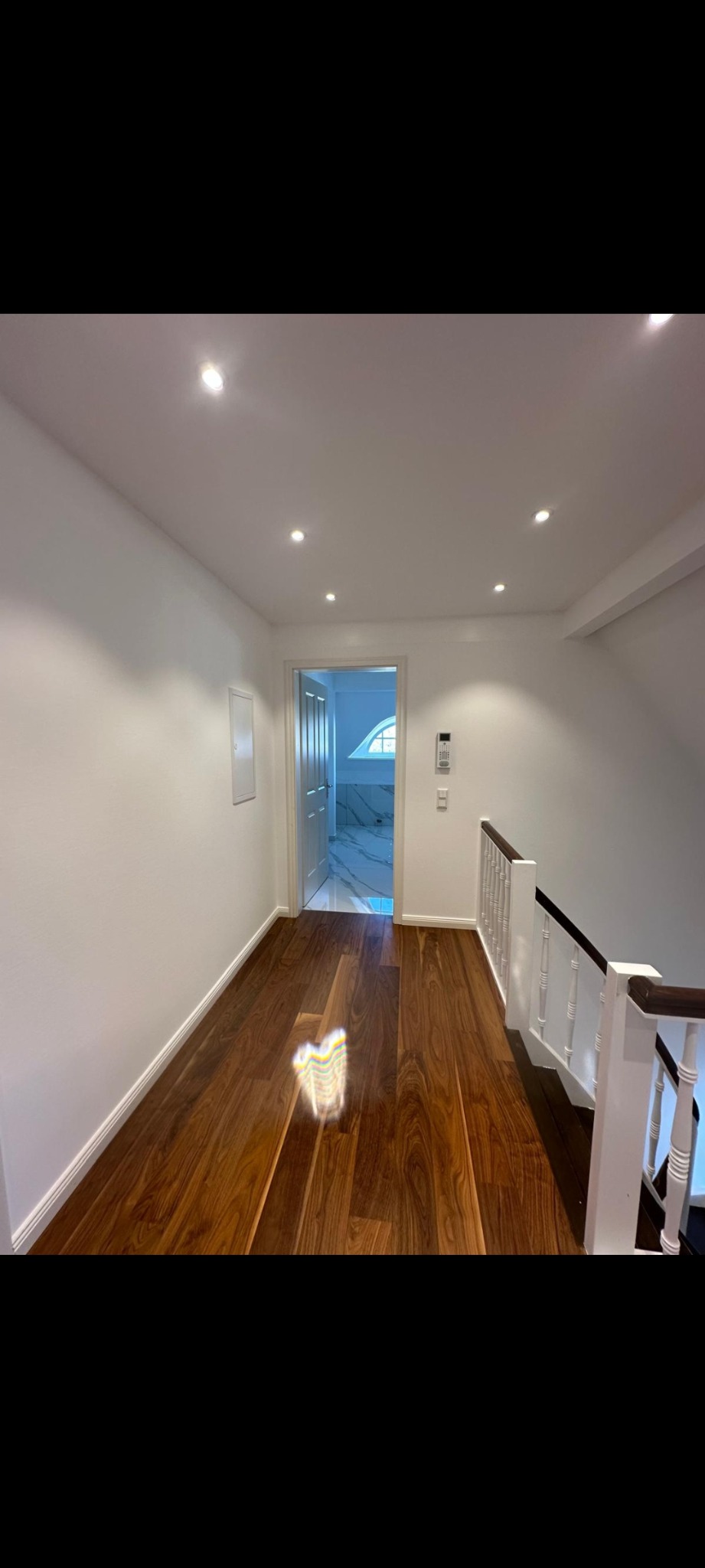 Korytarz na poddaszu z drewnianą podłogą i białymi ścianami. Widoczne schody z białą balustradą i wejście do łazienki z oknem dachowym.