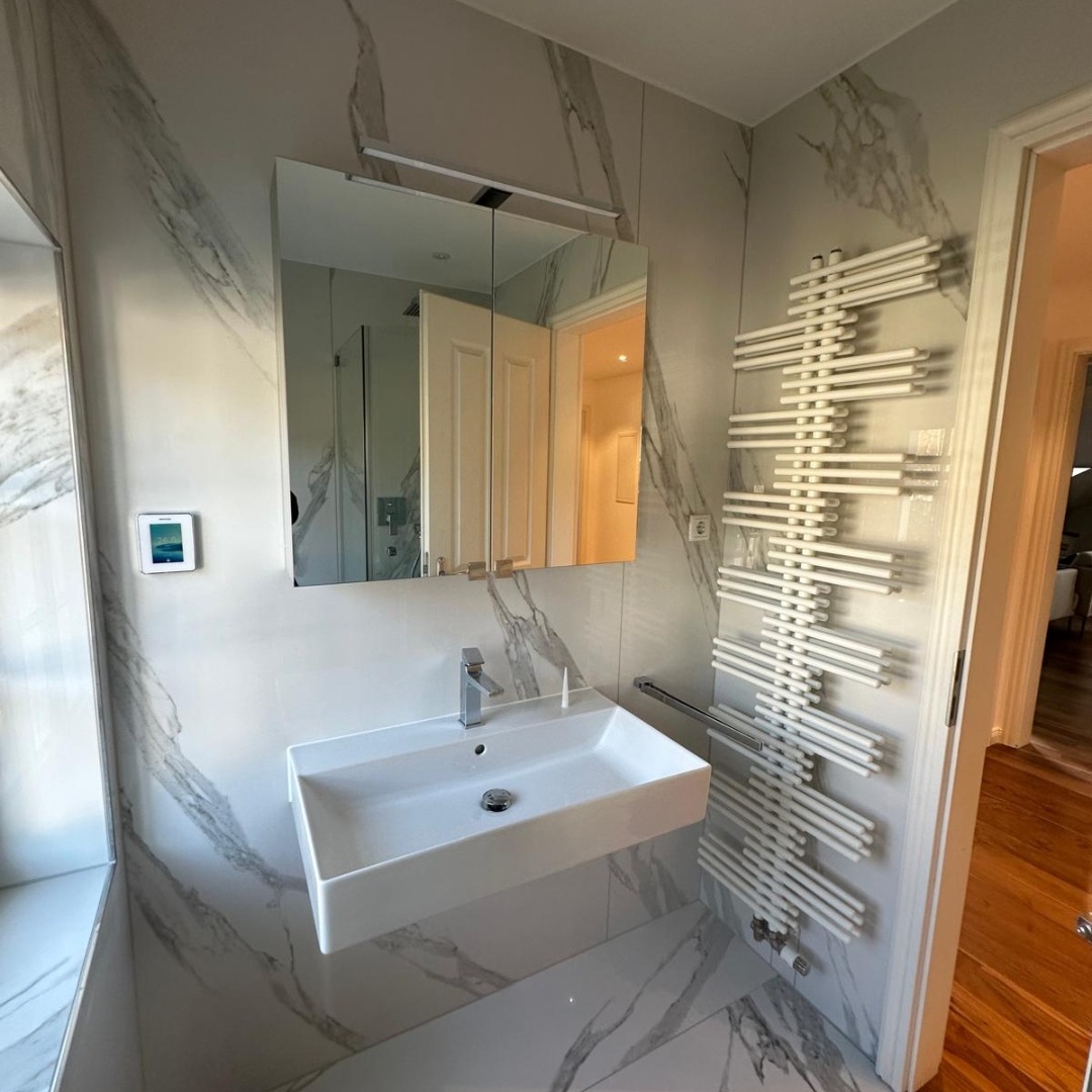 Nowoczesna łazienka z marmurowymi płytkami, wiszącą umywalką i grzejnikiem drabinkowym. Lustro z oświetleniem nad umywalką. Widok na wnętrze przez otwarte drzwi.