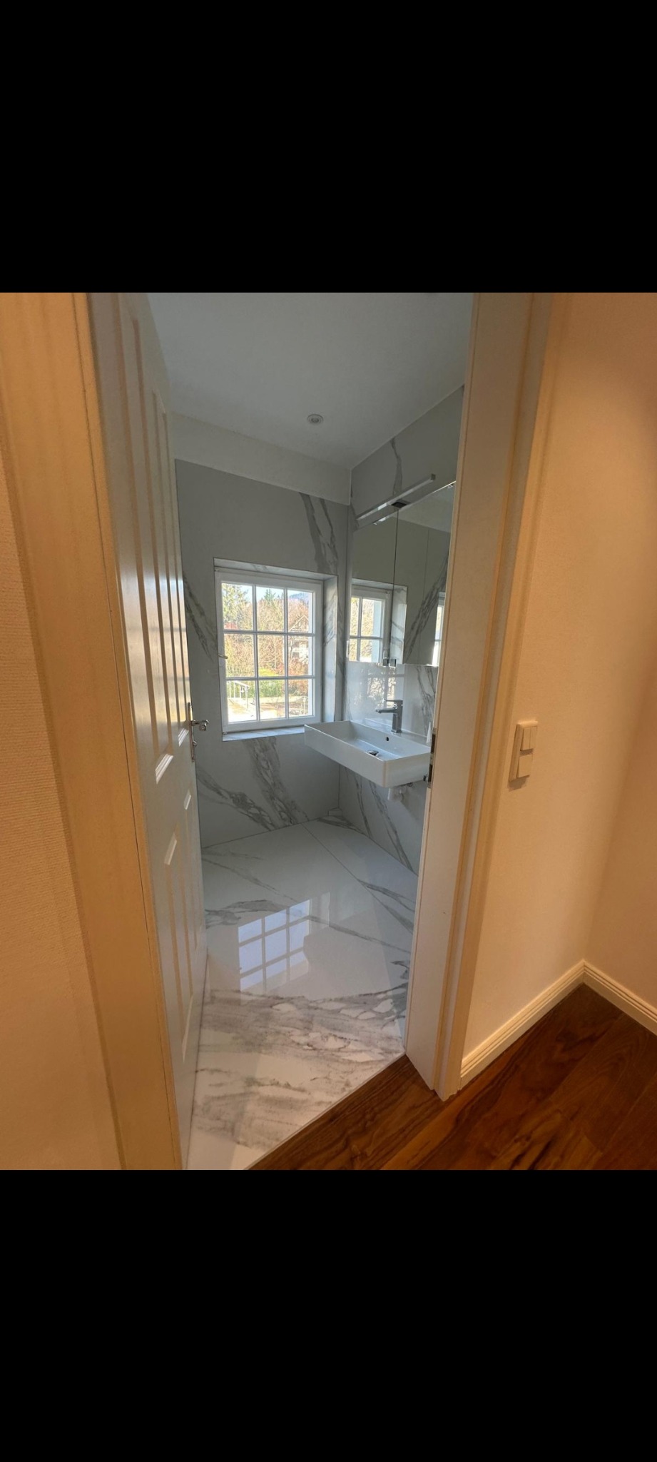 Wykończona łazienka z marmurowymi płytkami, widoczna z korytarza. Biała umywalka, okno z widokiem na zieleń. Podłoga z połyskiem odbija światło.