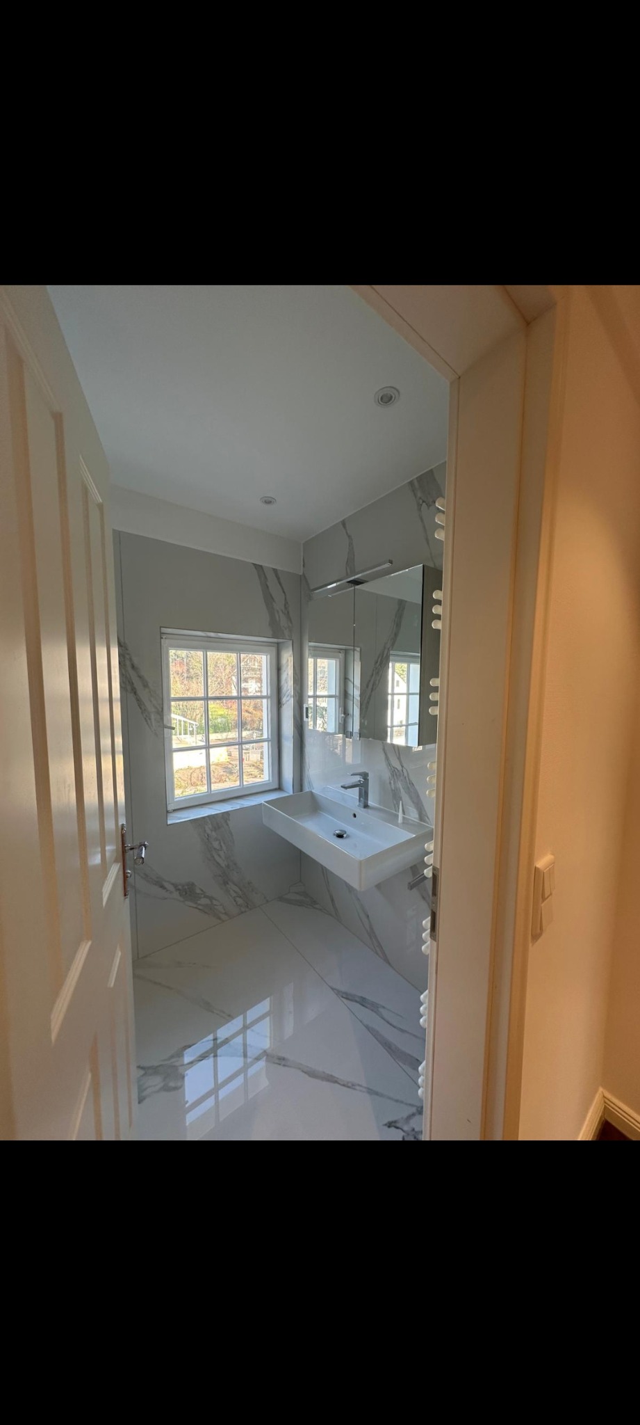 Nowoczesna łazienka z białymi płytkami imitującymi marmur, wisząca umywalka i lustro z oświetleniem. Widok zza uchylonych drzwi.