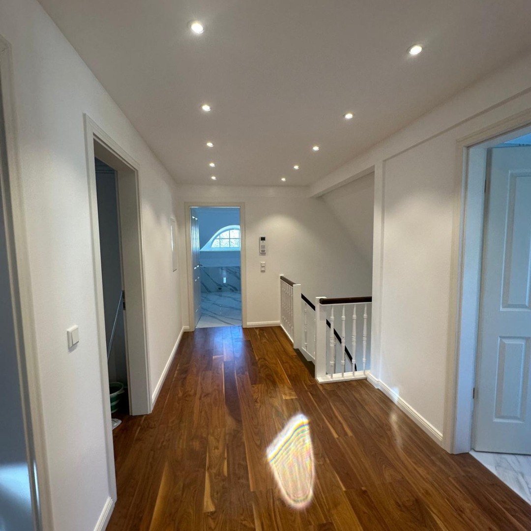 Korytarz z drewnianą podłogą i białymi ścianami. Widoczne drzwi do łazienki z marmurową podłogą i schody z białą balustradą. Oświetlenie punktowe w suficie.