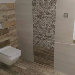 Wykończona łazienka na poddaszu z drewnopodobną glazurą na podłodze i ścianach oraz dekoracyjnym pasem płytek z ornamentami. Wisząca toaleta.
