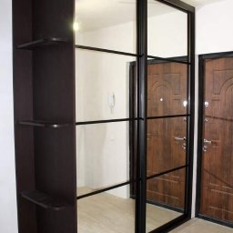 Usługi ogólnobudowlane Grzegorz Piotrzkowski - Wbudowana szafa z przesuwnymi drzwiami lustrzanymi, z ciemnobrązowymi ramami i półkami bocznymi, widoczna na tle wejścia z dwojgiem drewnianych drzwi.