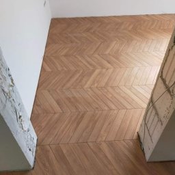 Usługi ogólnobudowlane Grzegorz Piotrzkowski - Ułożona podłoga z paneli drewnopodobnych w jodełkę, widoczne surowe wykończenie ścian i wystający fragment kolumny w stanie deweloperskim.