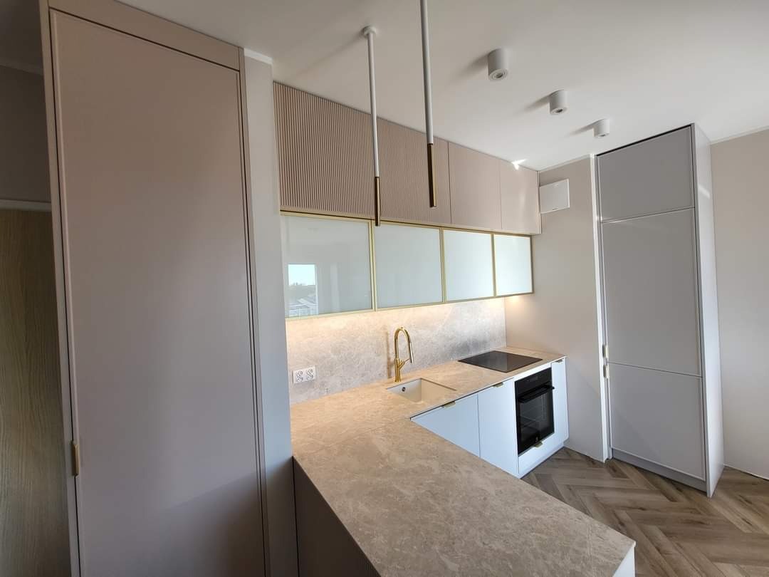 Nowoczesna kuchnia w odcieniach beżu i bieli z marmurowym blatem, złotą armaturą i oświetleniem punktowym, parkiet w jodełkę na podłodze.