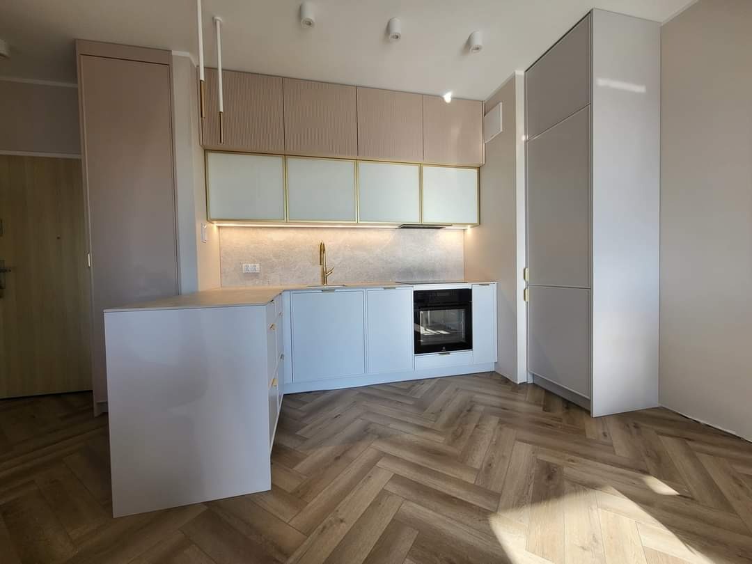 Nowoczesna kuchnia z białymi szafkami, złotymi akcentami i drewnianą podłogą ułożoną w jodełkę, oświetlona światłem LED pod szafkami wiszącymi.