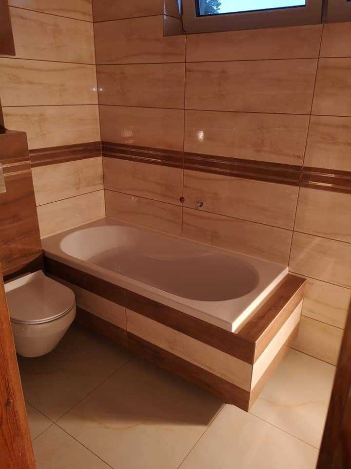 Jasna łazienka z wanną obudowaną drewnopodobnymi panelami, beżowe płytki na ścianach i podłodze, wisząca toaleta.