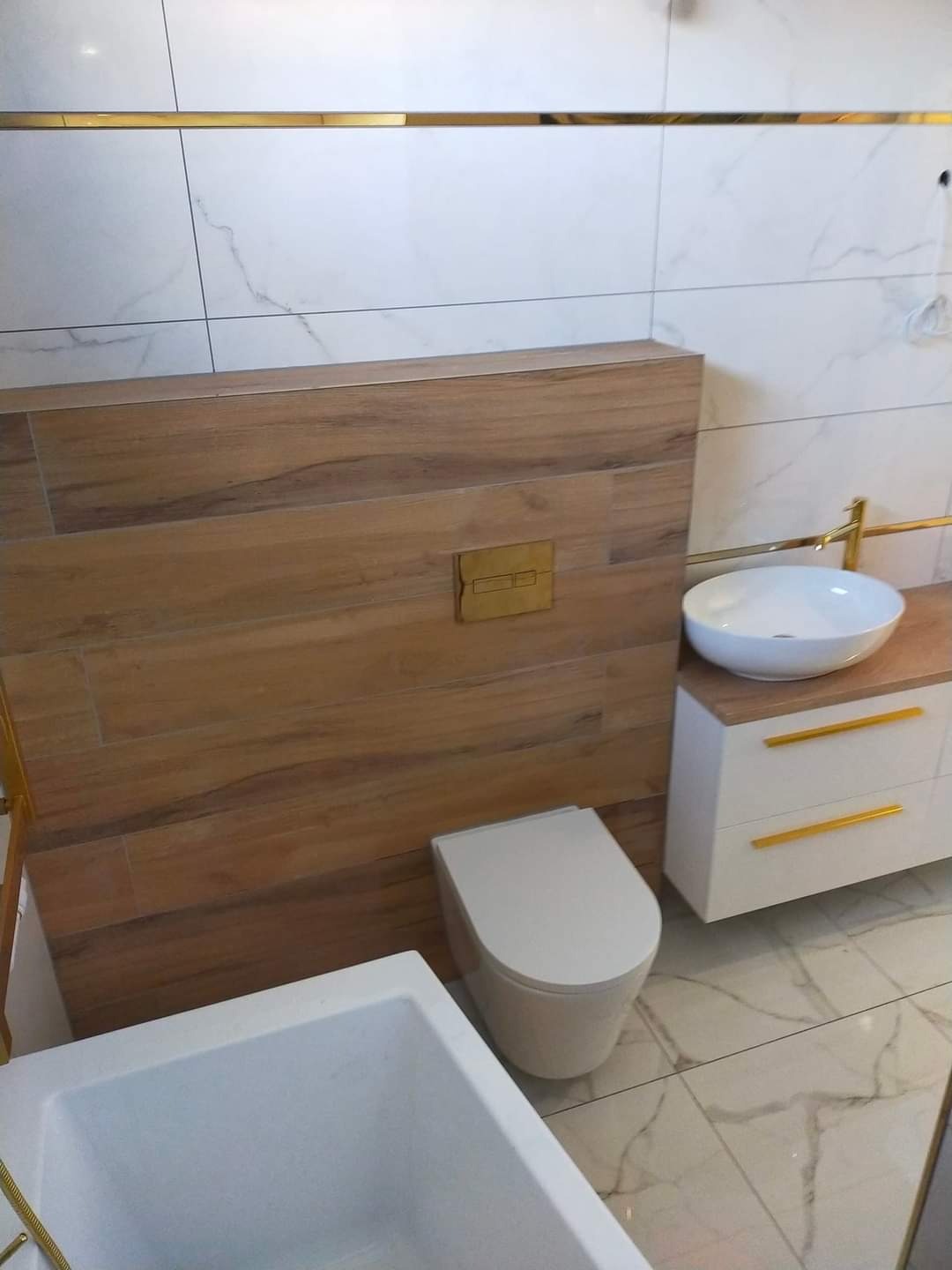 Nowoczesna łazienka w Gdyni po remoncie: białe płytki imitujące marmur, drewnopodobna zabudowa WC z mosiężnym przyciskiem, biała umywalka nablatowa z mosiężną armaturą, biała szafka pod umywalką...