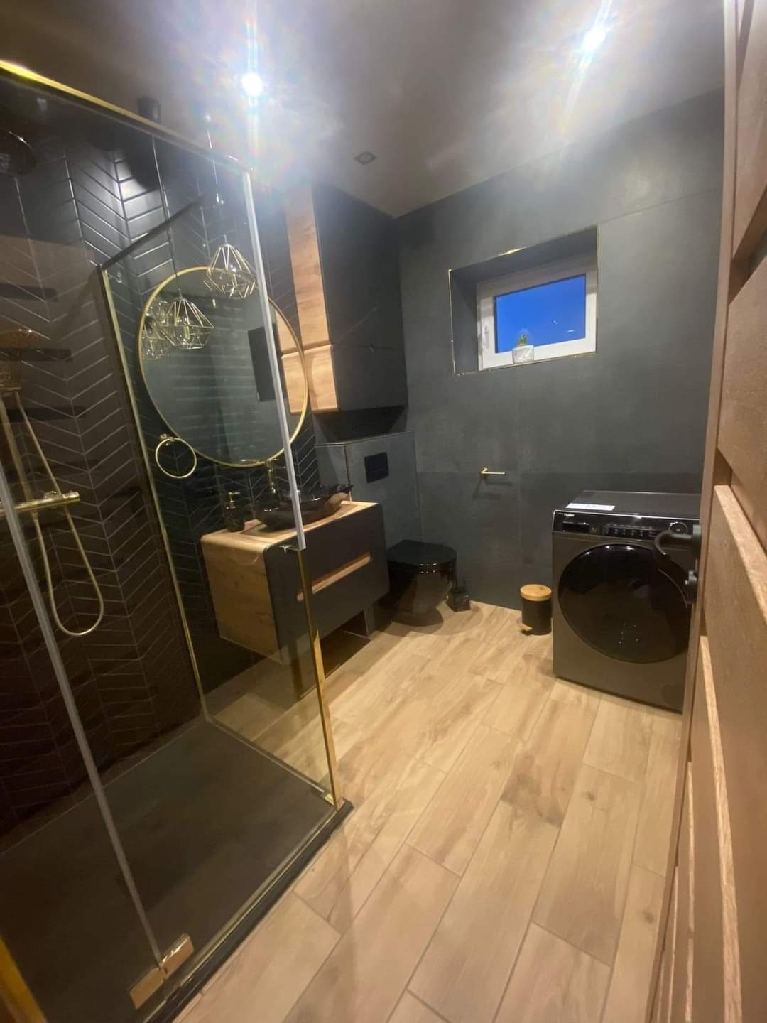 Nowoczesna łazienka w ciemnej tonacji z akcentami złota: kabina prysznicowa typu walk-in z czarną armaturą, umywalka nablatowa na szafce z drewnianymi frontami, pralka w rogu i płytki imitujące...