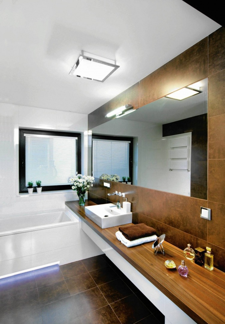 Nowoczesna łazienka z dużym lustrem, brązowymi płytkami i drewnianym blatem, biała umywalka i bukiet białych kwiatów na blacie.