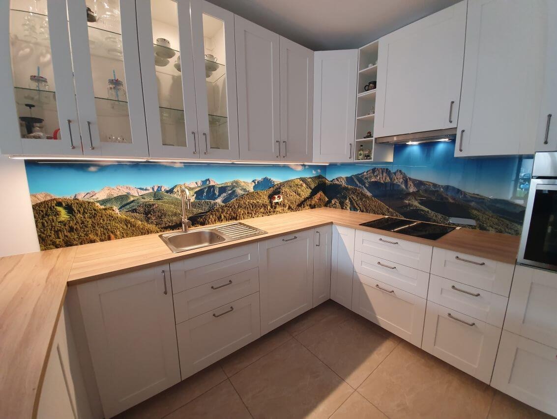 Biała kuchnia z górskim motywem na szklanym panelu nad blatem, widoczne szafki z przeszklonymi drzwiczkami i oświetleniem LED.