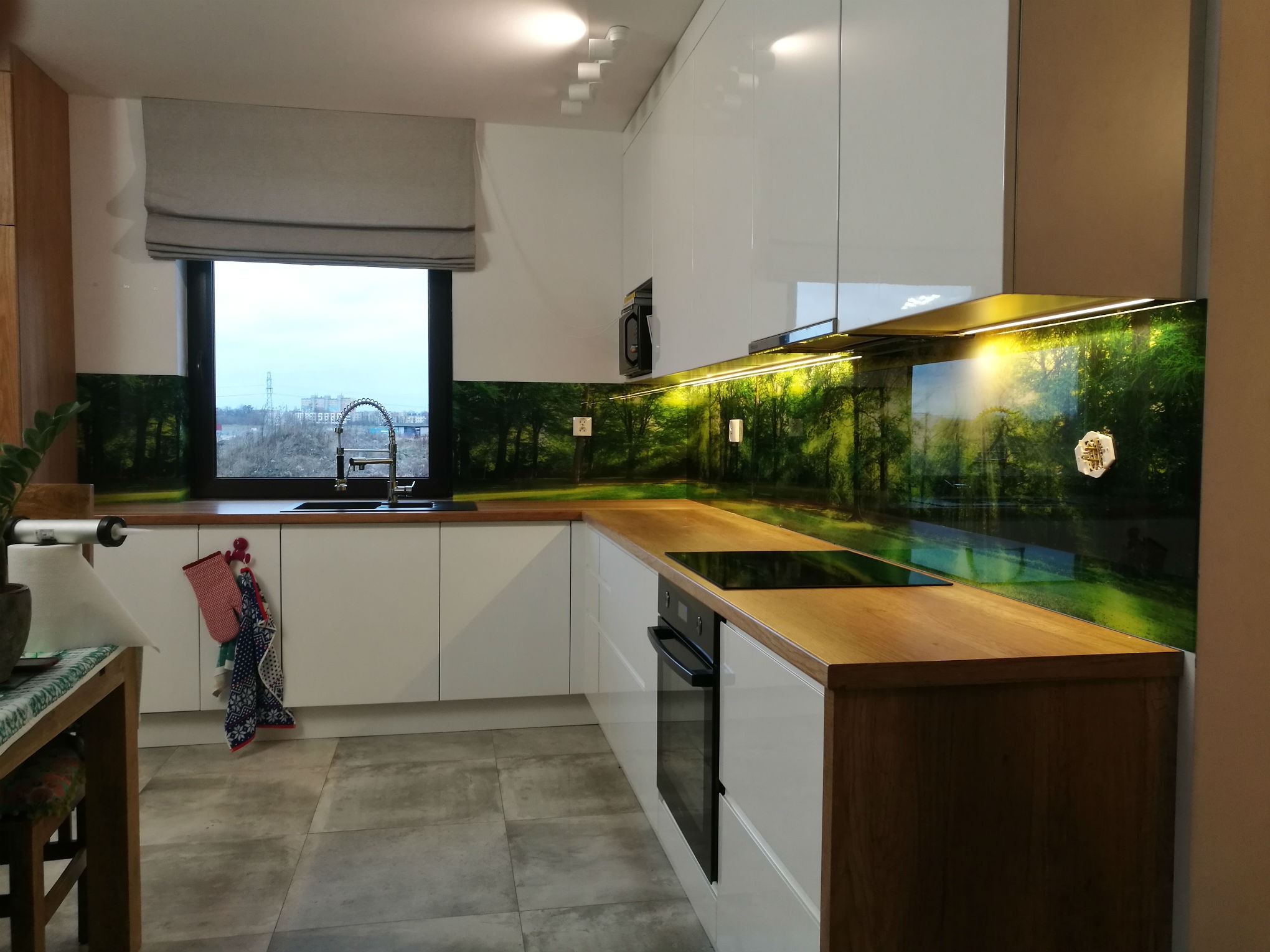 Nowoczesna kuchnia z białymi szafkami, drewnianym blatem i szklanym panelem ściennym z motywem leśnym, oświetlonym od spodu.