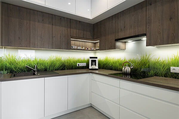 Nowoczesna kuchnia z białymi szafkami i drewnianymi górnymi szafkami, podświetlony panel szklany z motywem trawy na ścianie, srebrny czajnik i ekspres do kawy na blacie.