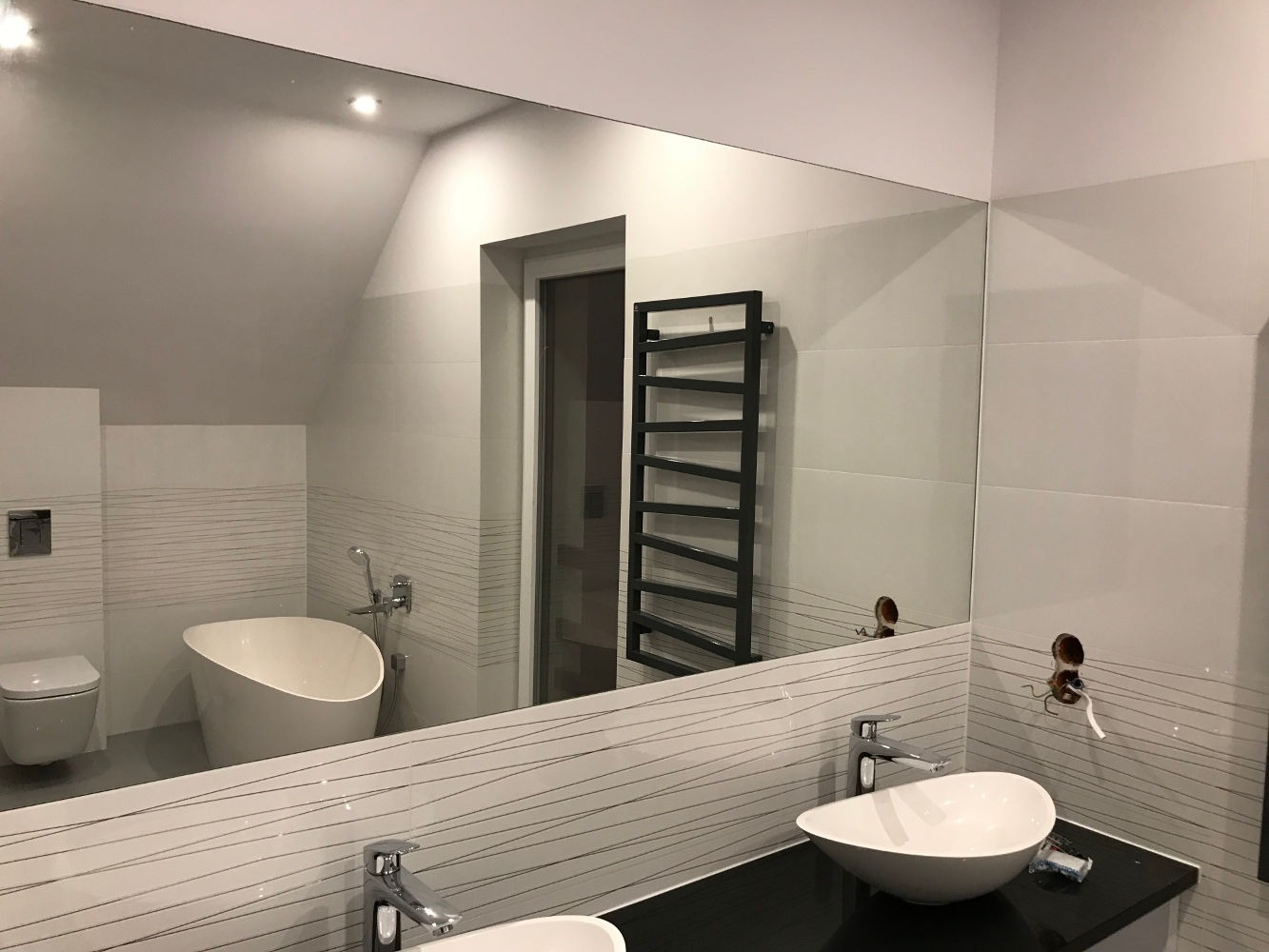 Duże lustro w łazience z widocznym odbiciem wanny, toalety i grzejnika drabinkowego; w tle białe płytki z szarymi liniami, widoczne niedokończone podłączenie baterii umywalkowej.