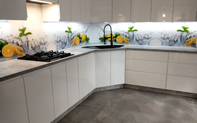 Biała, nowoczesna kuchnia z błyszczącymi frontami, szarym blatem i panelem szklanym nad blatem z motywem cytryn i rozpryskującej się wody, czarna armatura i płyta grzewcza.