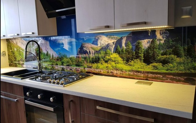 Szklany panel ścienny w kuchni z motywem górskiego krajobrazu, widoczny zlew, płyta gazowa i fragment piekarnika.