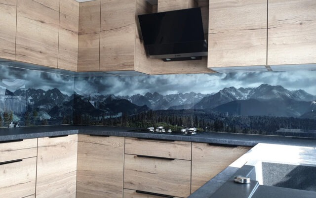 Szklany panel kuchenny z motywem górskim, drewniane szafki w jasnym odcieniu, czarny okap nad płytą grzewczą.