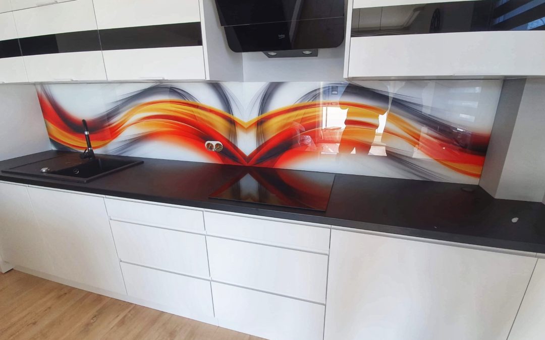 Szklany panel dekoracyjny z abstrakcyjnym wzorem w odcieniach czerwieni, pomarańczy i szarości, zamontowany nad czarnym blatem kuchennym i białymi szafkami, z widocznym gniazdkiem elektrycznym.