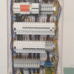 Otwarta skrzynka elektryczna z widocznymi bezpiecznikami, przewodami o różnych kolorach i elementami montażowymi, zamontowana na białej ścianie.