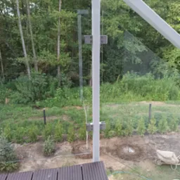 Szklana balustrada z białymi słupkami i mocowaniami na tarasie z kompozytowych desek, widok na ogród z posadzonymi drzewkami