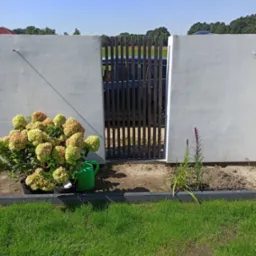 Nowoczesna brama wjazdowa z pionowych metalowych profili, wkomponowana w biały, betonowy mur ogrodzeniowy, z widocznym samochodem w tle; z przodu rabata z hortensjami w donicach i konewką.