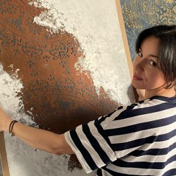 EMDekor - Dekoracyjna ściana z efektem stiuku weneckiego i wzorem, prezentowana przez kobietę w bluzie w paski. Tło ściany z ornamentami. Unikalna faktura i kolorystyka.