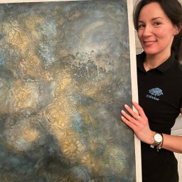 EMDekor - Dekoracyjna ściana z efektem tynku strukturalnego w odcieniach złota i błękitu, prezentowana przez uśmiechniętą kobietę w koszulce z logo firmy.