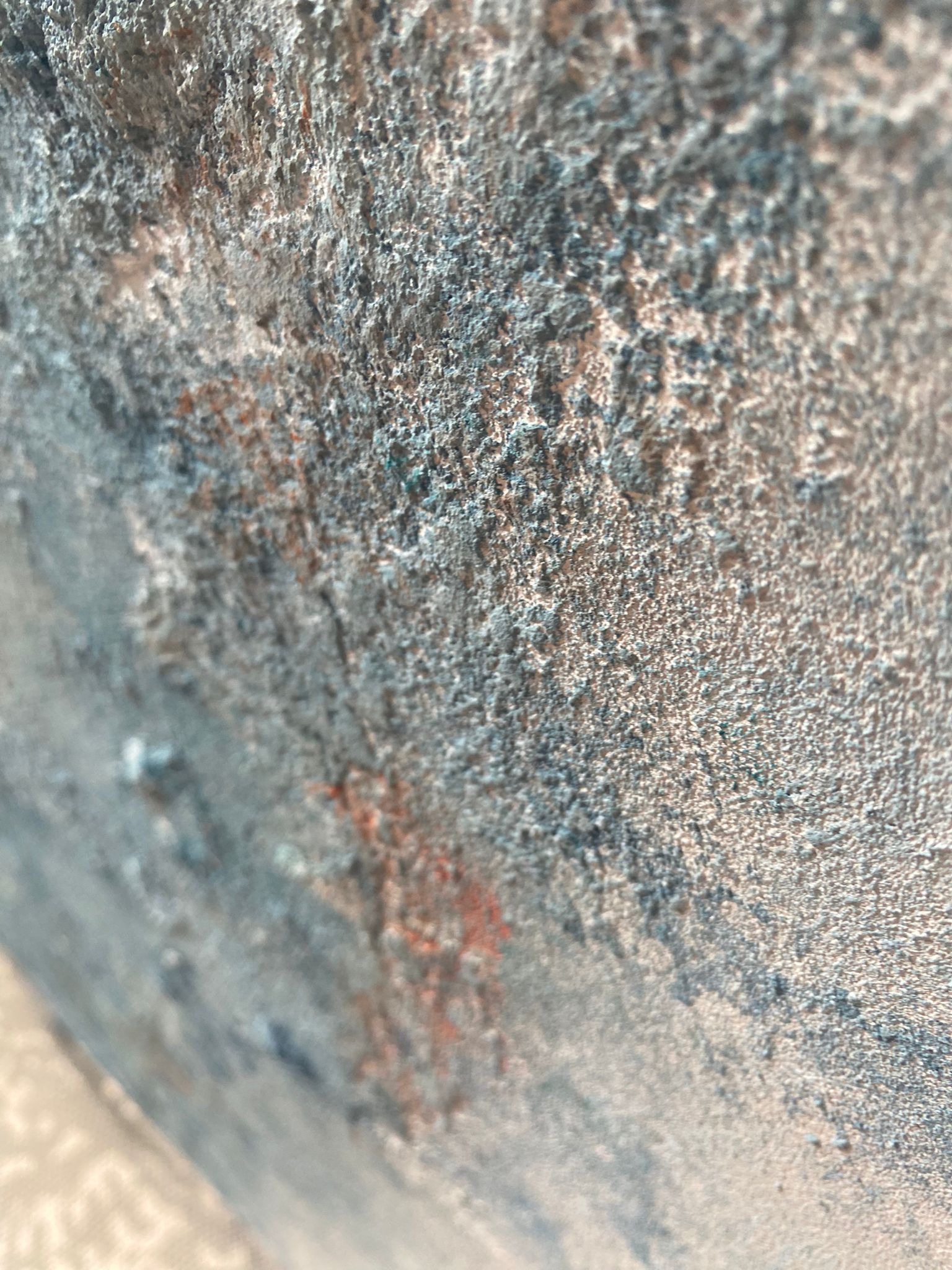 Szara, chropowata powierzchnia tynku dekoracyjnego z delikatnymi, rdzawymi akcentami, sfotografowana pod kątem, ukazująca fakturę i nierówności.