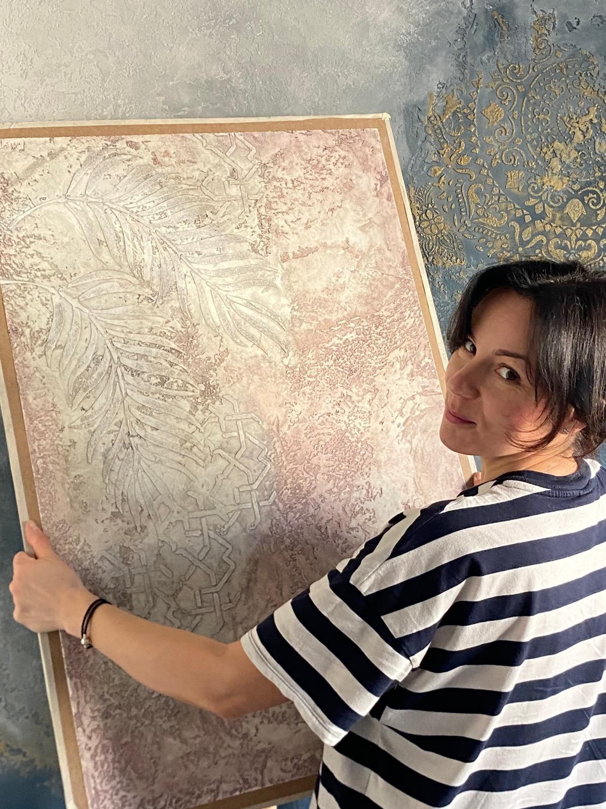 Kobieta prezentuje dekoracyjny panel ścienny z efektem tynku strukturalnego i motywem liści palmowych. W tle ściana z dekoracyjnym tynkiem w odcieniach niebieskiego i złota.