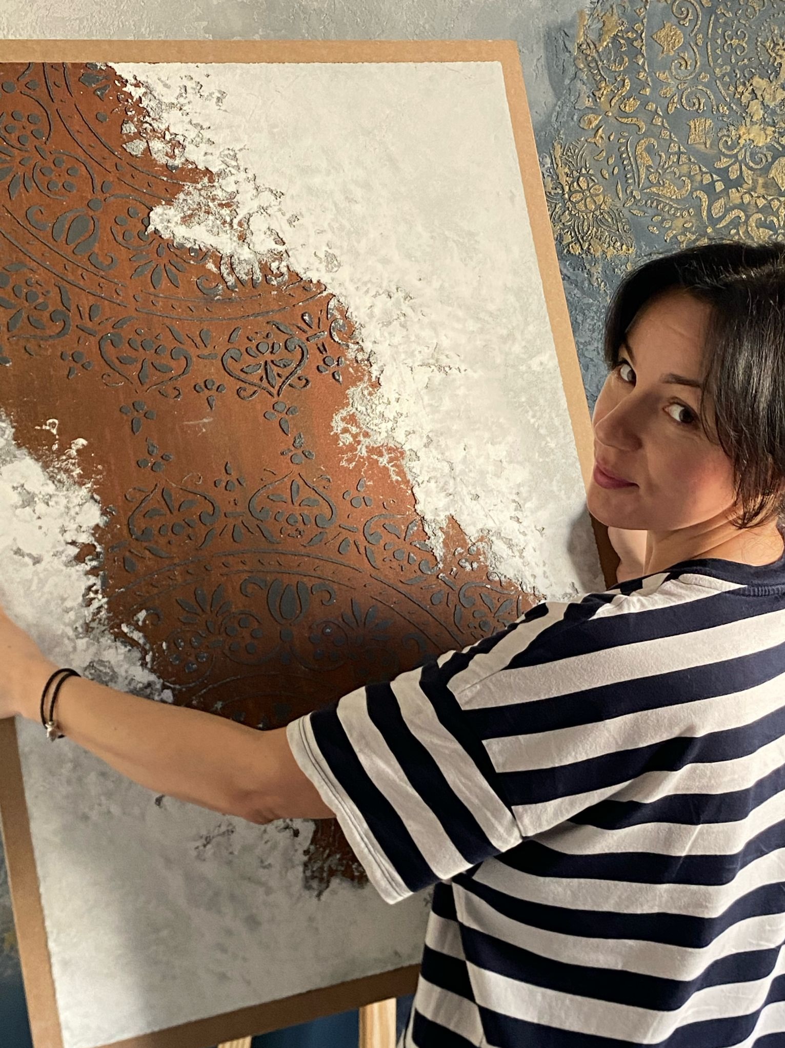 Dekoracyjna ściana z efektem stiuku weneckiego i wzorem, prezentowana przez kobietę w bluzie w paski. Tło ściany z ornamentami. Unikalna faktura i kolorystyka.